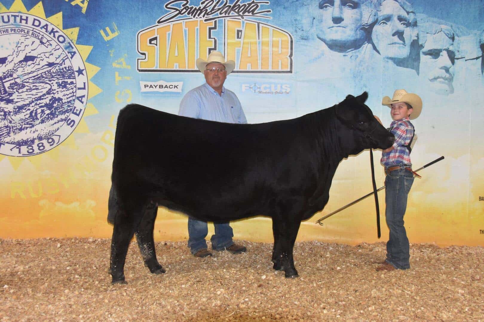 A man and a child in cowboy hats stand next to a black cow at the South Dakota State Fair, with a backdrop featuring Mount Rushmore and fair logos. The child holds the cow by a lead.