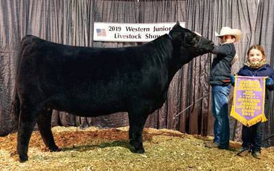 A boy in a cowboy hat stands beside a large black cow at the 2019 Western Junior Livestock Show. A girl next to them holds a purple and yellow champion banner.
