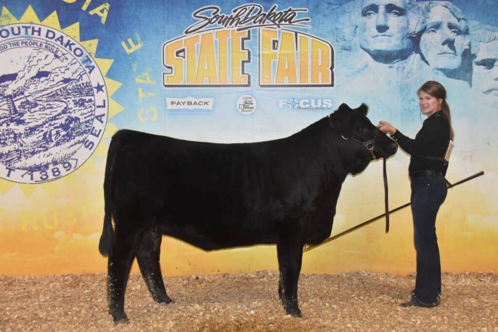 A woman holds a black cow on a lead at the South Dakota State Fair, posing in front of a backdrop featuring the state seal and Mount Rushmore.