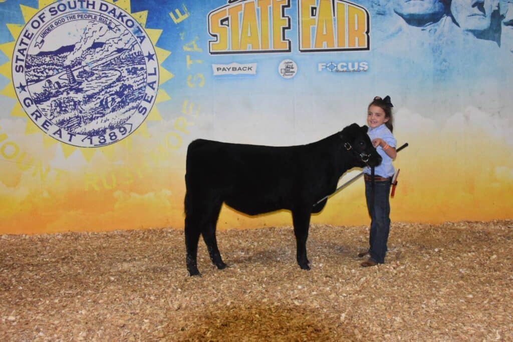 A young girl stands smiling next to a black calf at the South Dakota State Fair, holding its halter in a livestock show ring with wood shavings on the ground and a large state seal backdrop behind them.