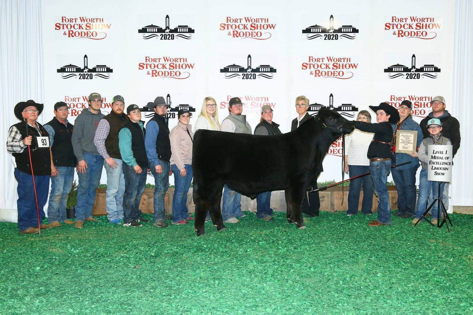 A group of people stand behind a black steer at the Fort Worth Stock Show & Rodeo, posing for a photo with banners and awards on a green floor in front of a backdrop with event logos.