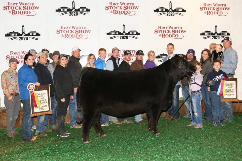 A large group of people stands around a black steer holding banners and ribbons at the Fort Worth Stock Show & Rodeo 2020, posing for a photo in front of a branded backdrop.