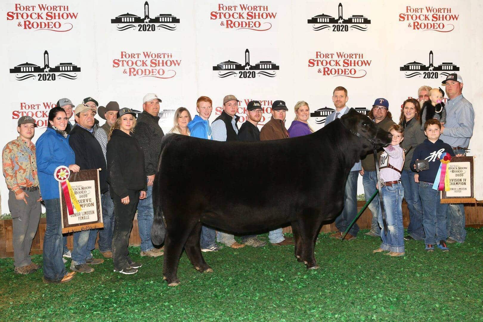 A large group of people stands around a black steer holding banners and ribbons at the Fort Worth Stock Show & Rodeo 2020, posing for a photo in front of a branded backdrop.