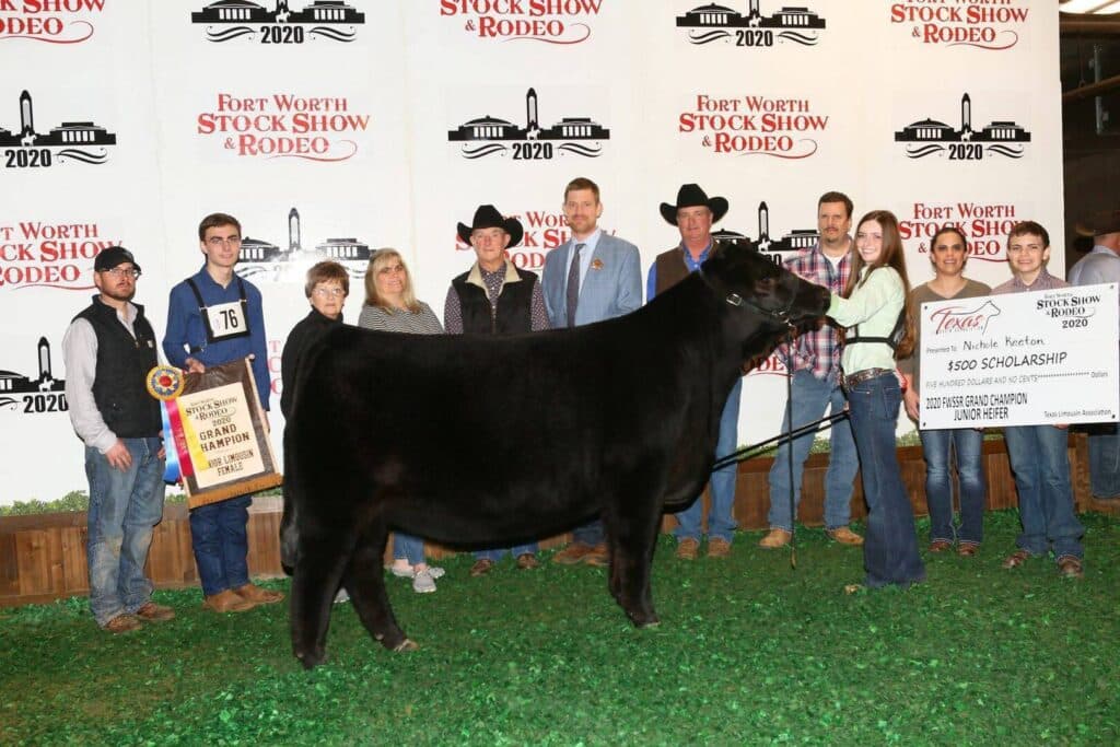 A group of people pose indoors with a prize-winning black steer at the 2020 Fort Worth Stock Show & Rodeo. They display awards, including a ribbon and a large scholarship check.