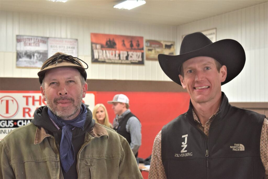 Two men pose indoors, both wearing jackets; one has a flat cap and scarf, the other a black cowboy hat and branded vest. People and Western-themed signs are visible in the background.