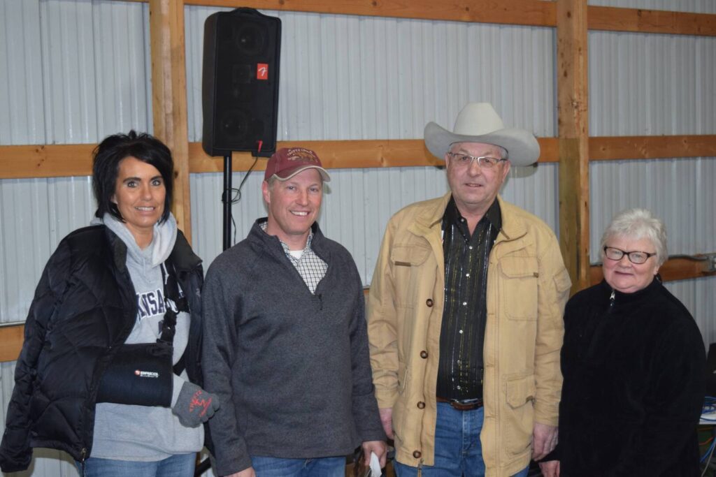 Four adults stand indoors in front of a corrugated metal wall and wooden beams. One woman wears a puffy jacket and arm sling, two men wear jackets and hats, and another woman wears glasses and a black jacket.