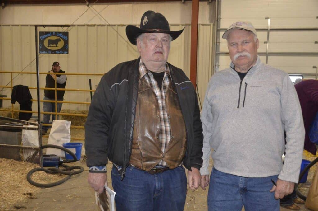 Two older men stand indoors on straw-covered ground; one wears a cowboy hat, plaid shirt, and leather vest, while the other wears a light pullover and cap. Animal pens and a “Animal Calves” sign are visible in the background.
