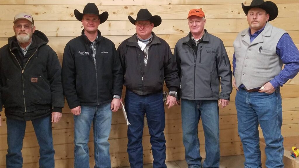 Five men wearing jackets and cowboy hats stand in a row against a wooden wall, posing for the photo. Four of the men wear black hats, and one wears an orange cap.
