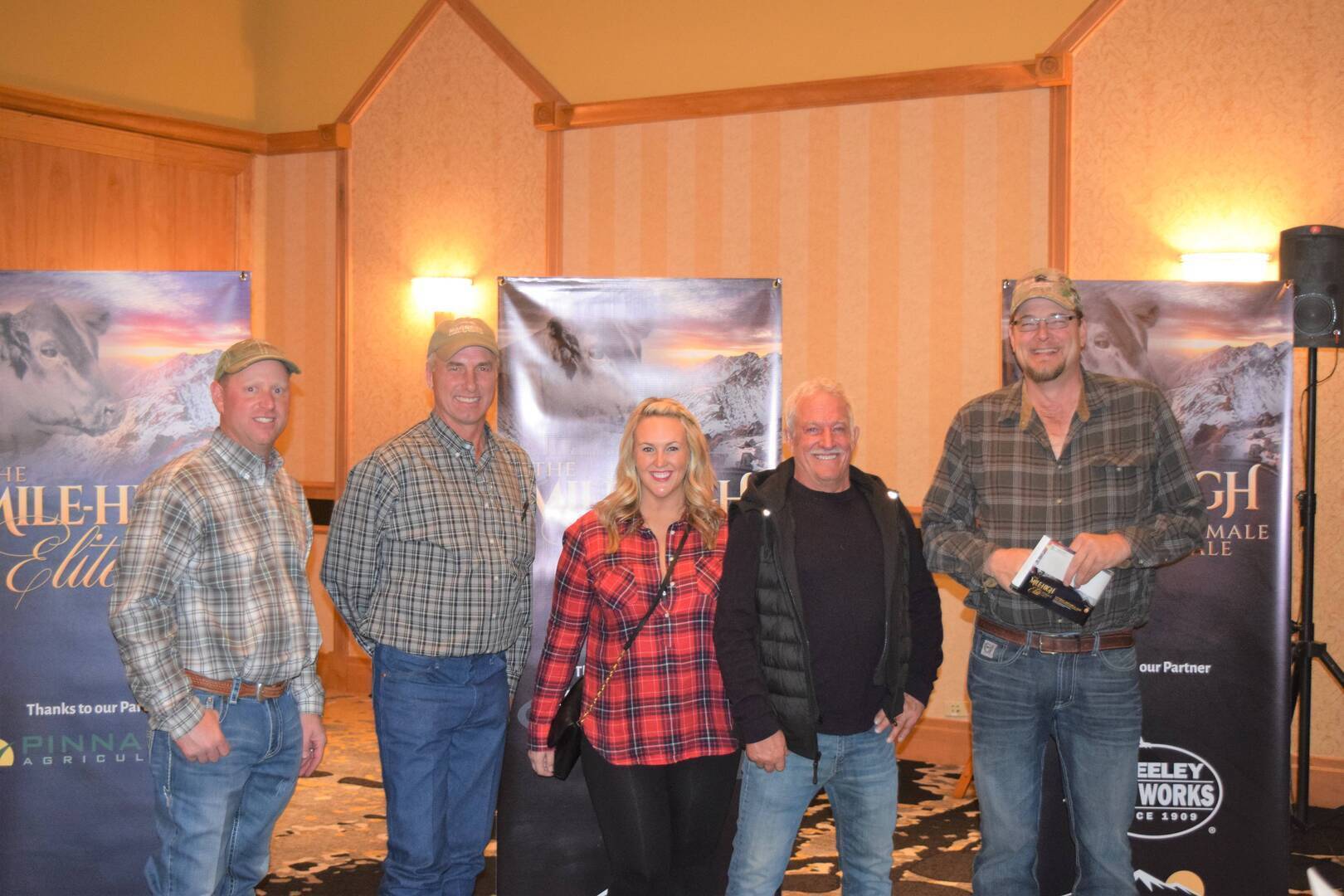 Five people stand indoors at an event, four men and one woman, all dressed in casual clothing and plaid shirts. Banners and wooden walls are visible in the background. The group is smiling at the camera.