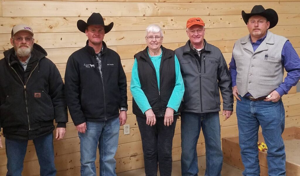 Five adults stand indoors against a wooden wall. Four men wear cowboy hats or caps and casual jackets; one woman stands in the center, smiling, wearing glasses and a turquoise shirt with a black vest.