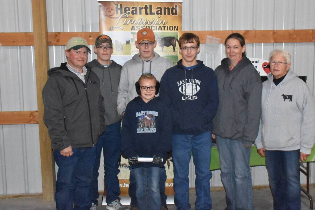 Seven people stand indoors in front of a Heartland Limousin Association banner, wearing casual clothing and hoodies. The group appears to be at an agricultural or community event in a barn-like setting.