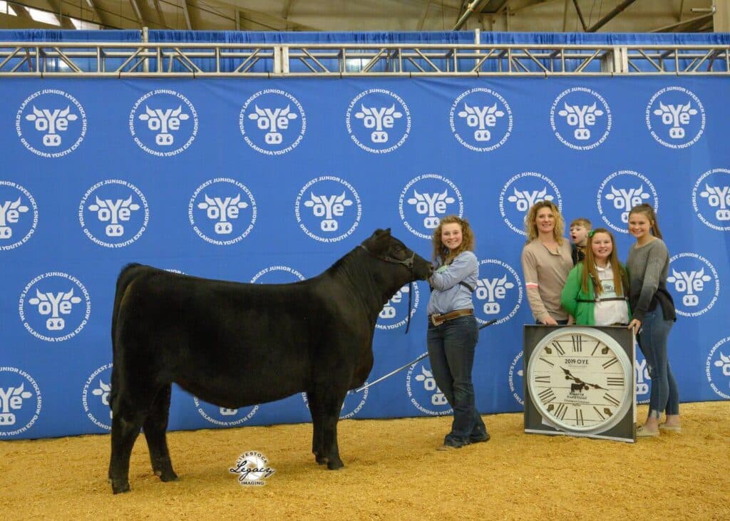 Five people pose with a black cow in front of a blue backdrop with white logos. One person holds the cow, while another holds a large clock with “OYE 2021” on it. Everyone is smiling.