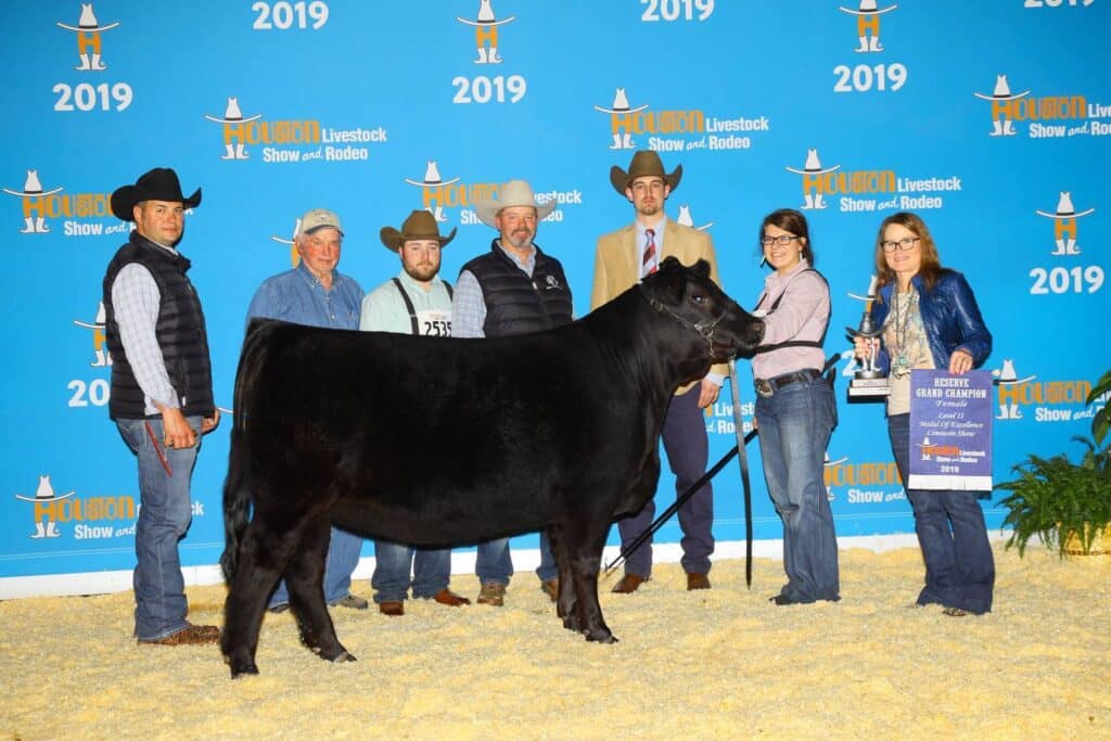 A group of people stand around a black cow at the 2019 Houston Livestock Show and Rodeo, posing for a photo in front of a blue event backdrop, with one person holding a trophy and another holding a plaque.
