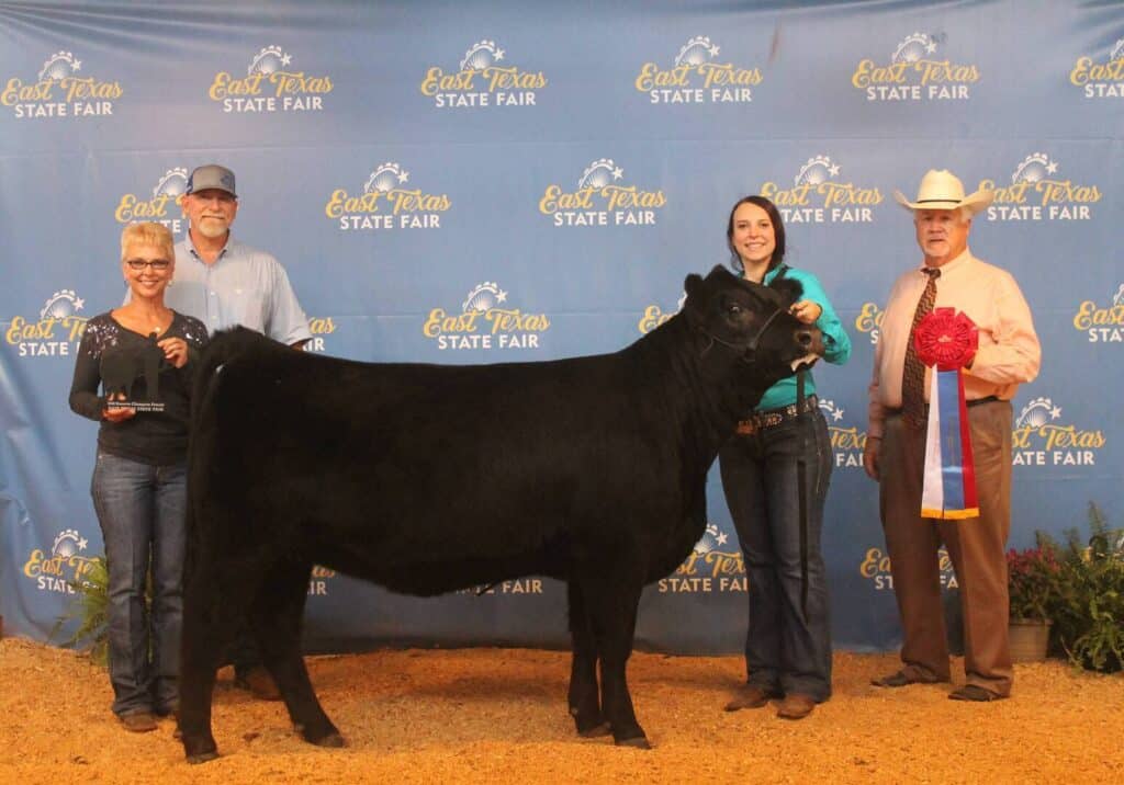 Four people pose with a black cow at the East Texas State Fair. Two women stand by the cow, one holding a trophy. Two men stand at either end, one holding a large ribbon. A blue fair banner is in the background.