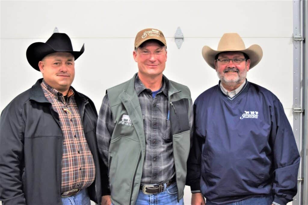 Three men wearing cowboy hats stand side by side, smiling at the camera. They are dressed in casual western-style clothing, including plaid shirts, jackets, and vests, with a plain light-colored wall behind them.