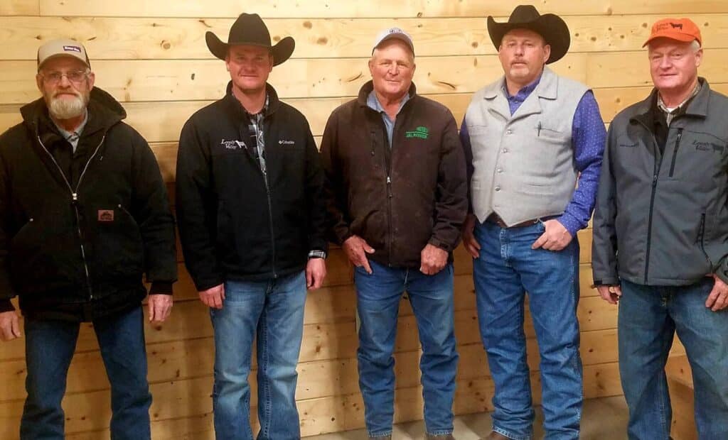 Five men stand side by side against a wooden wall. They are dressed in casual outdoor attire, including jackets, jeans, hats, and caps, with two wearing cowboy hats. They are facing the camera and appear to be posing for a group photo.