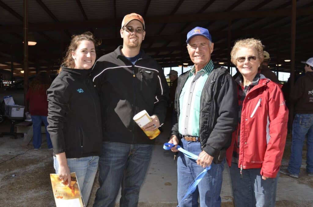 Four people stand together smiling at an outdoor event. One holds a magazine, another a container and paper. They are dressed in jackets and hats, with a large open barn structure and other people in the background.
