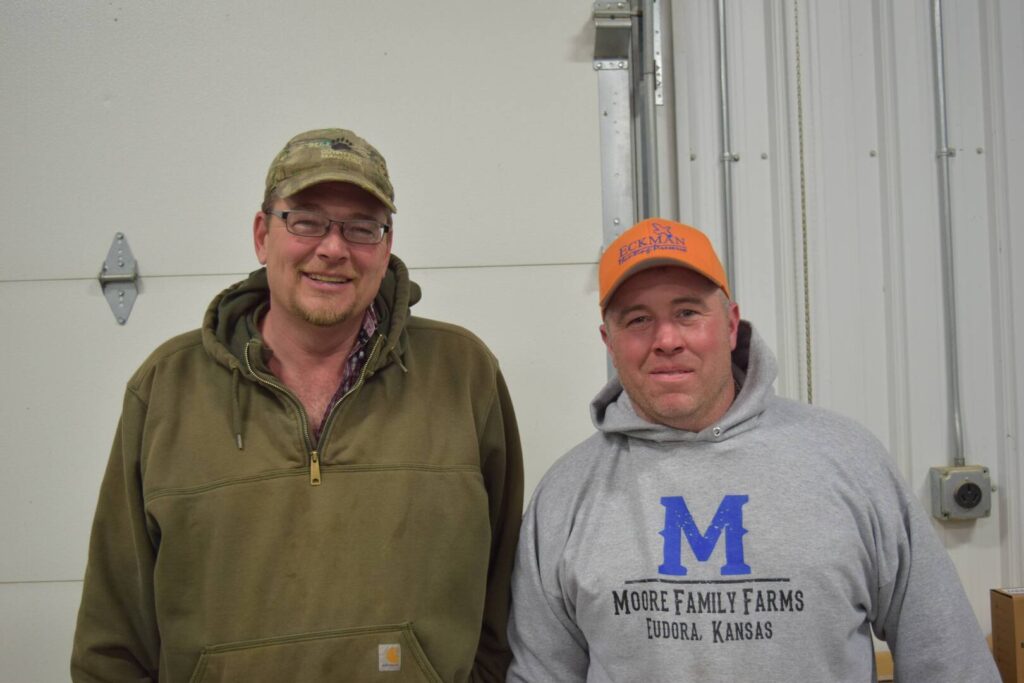 Two men standing indoors, both smiling. One wears glasses, a green hoodie, and a camouflage cap. The other wears a gray Moore Family Farms hoodie and an orange cap. They are in front of a white wall with metal details.