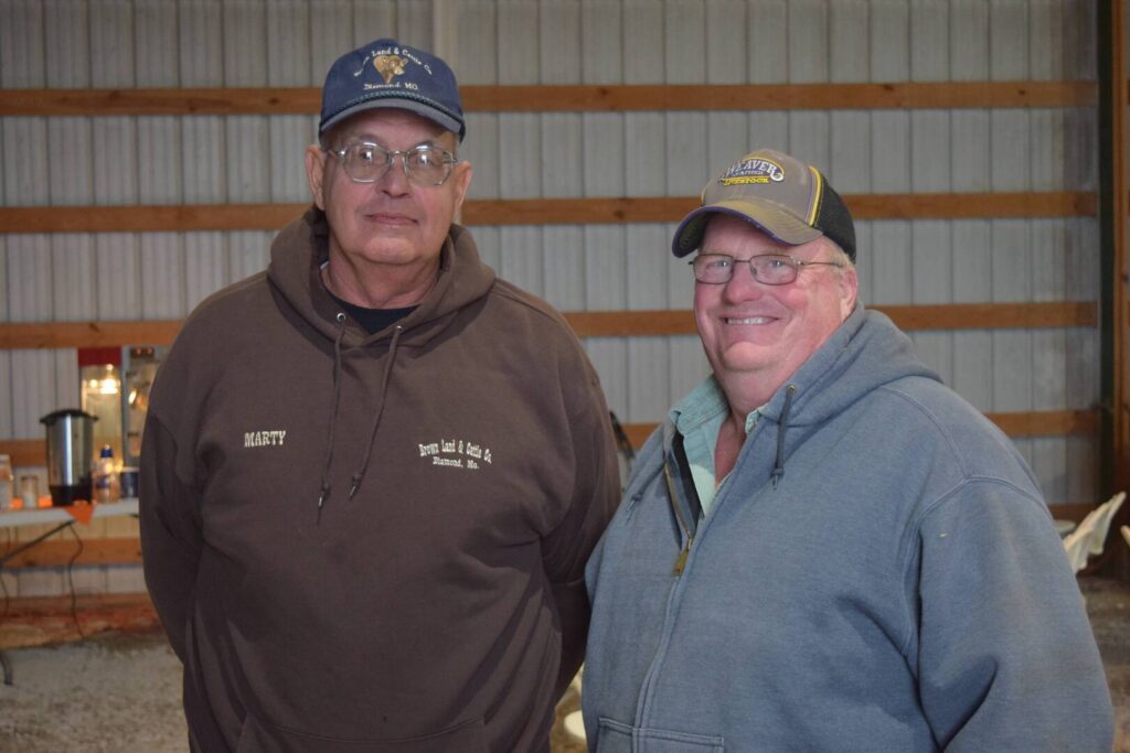 Two older men wearing glasses, baseball caps, and hooded sweatshirts stand and smile inside a metal-sided building. One sweatshirt is labeled Marty. The background shows a coffee maker on a table and wooden beams.
