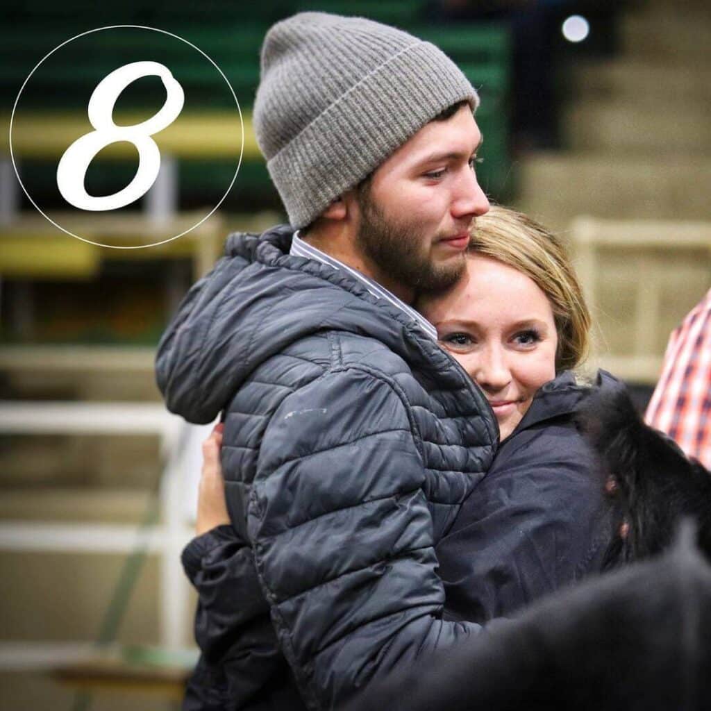 A young man in a gray beanie and jacket hugs a smiling woman in a black jacket. The number 8 is overlaid in white in the top left corner of the image. They are indoors, with blurred benches in the background.