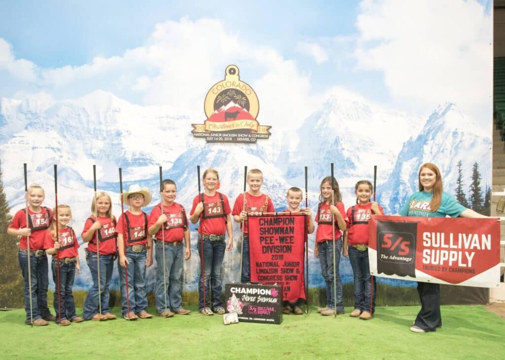 A group of children in red shirts and jeans stand in a row holding show sticks, smiling for a photo at a livestock show. An adult woman stands at the end holding a red Sullivan Supply banner. Mountains are pictured in the backdrop.