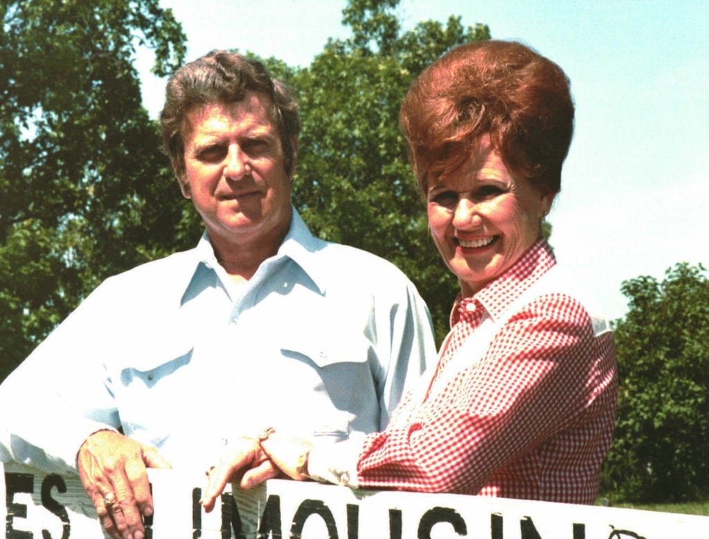 A man in a light blue shirt and a woman in a red checkered shirt stand outdoors in front of trees, smiling at the camera.