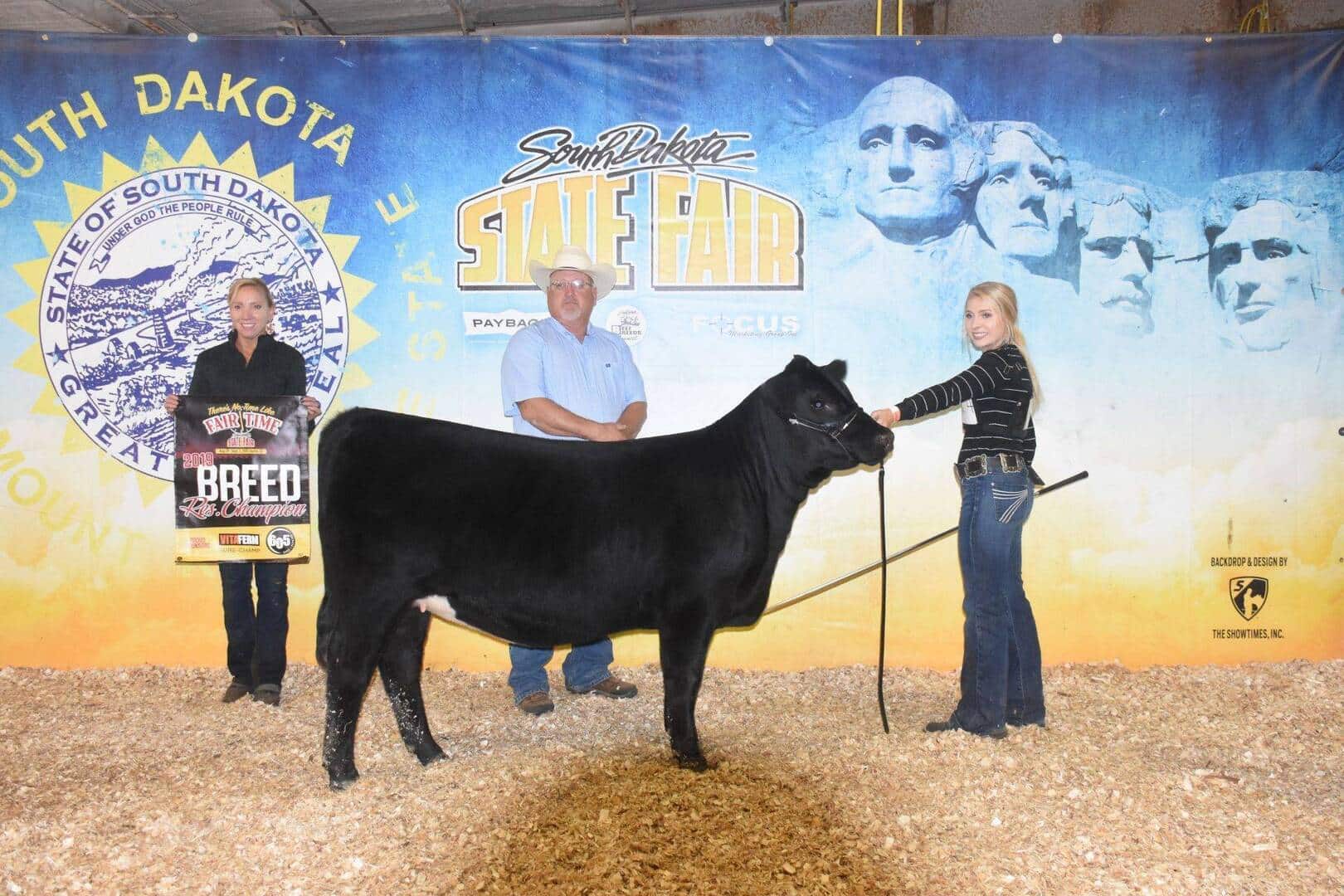Three people pose with a black cow at the South Dakota State Fair. One woman holds a Breed Champion sign, while another holds the cows lead. A man stands behind them with his hands clasped. The backdrop features Mount Rushmore.