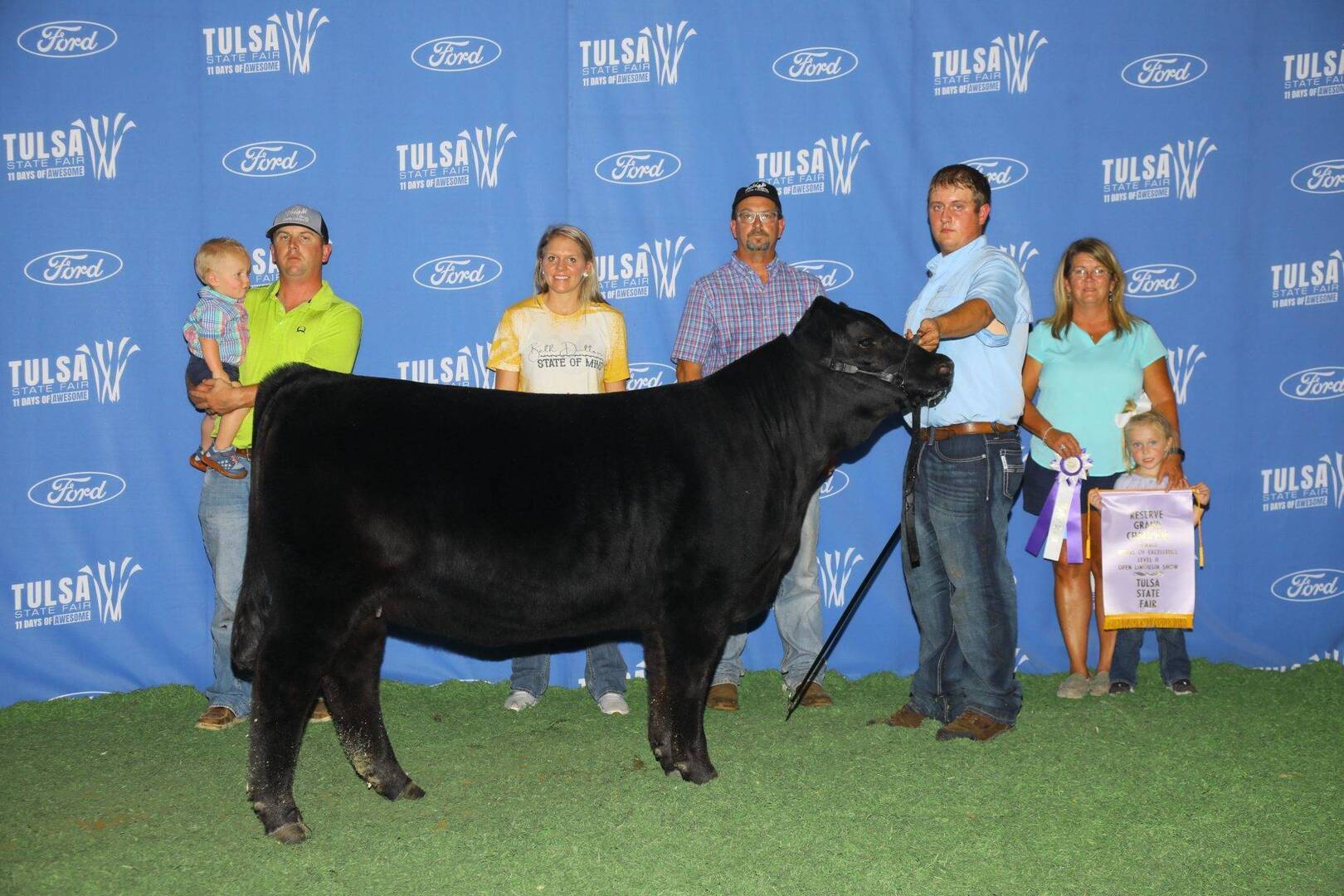 A group of people pose with a black cow at the Tulsa State Fair. One person holds the cow’s halter, while another holds a ribbon and banner. A blue backdrop with Tulsa State Fair and Ford logos is behind them.