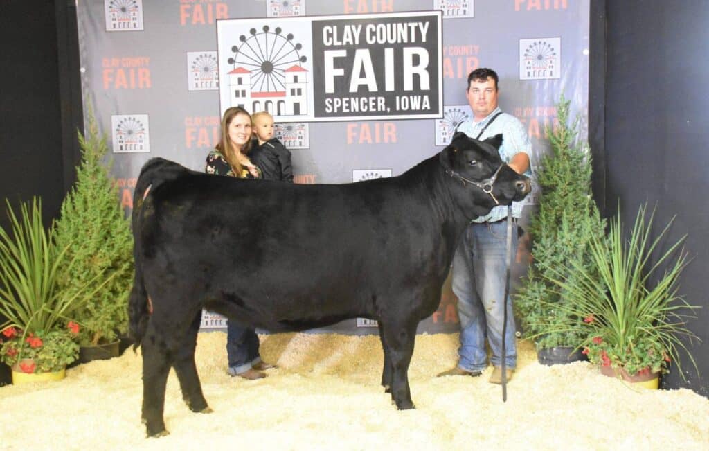 Three people pose with a large black steer in front of a backdrop that reads Clay County Fair Spencer, Iowa. The ground is covered with straw, and potted plants surround the group.