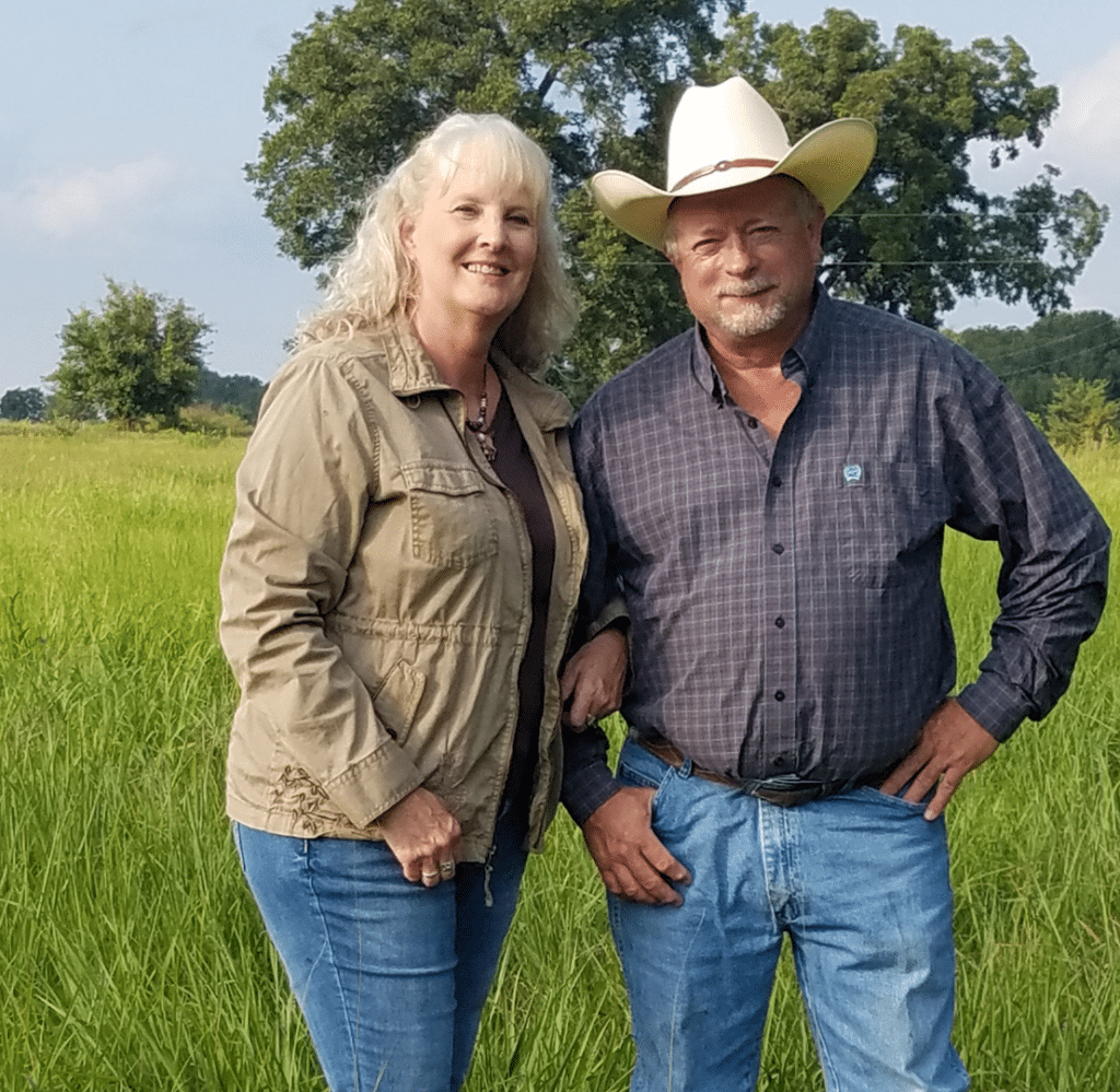 A woman and a man wearing a cowboy hat stand together in a grassy field on a sunny day, smiling at the camera with trees in the background.