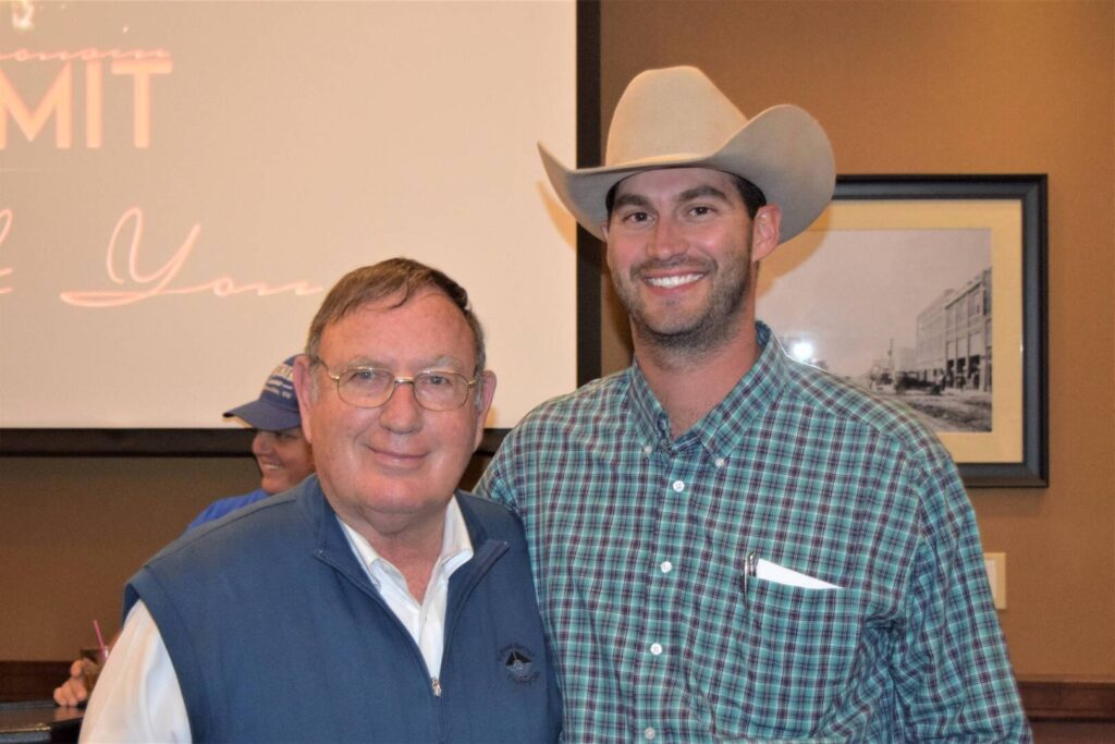 Two men posing and smiling indoors; one wears glasses and a blue vest, the other wears a cowboy hat and plaid shirt. A projected screen and a framed photo are in the background.