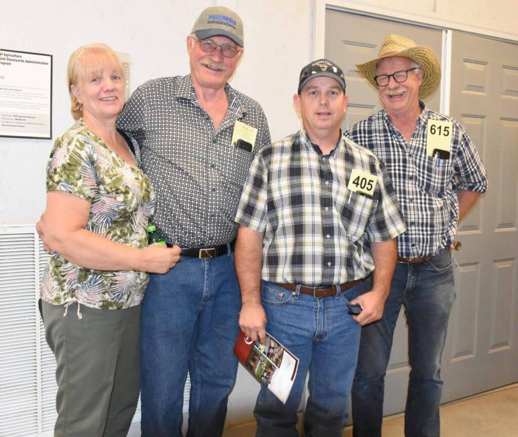 Four adults stand together indoors, smiling at the camera. Three men wear numbered tags, jeans, and hats; one woman in a floral shirt stands beside them. They appear to be attending an event or auction.