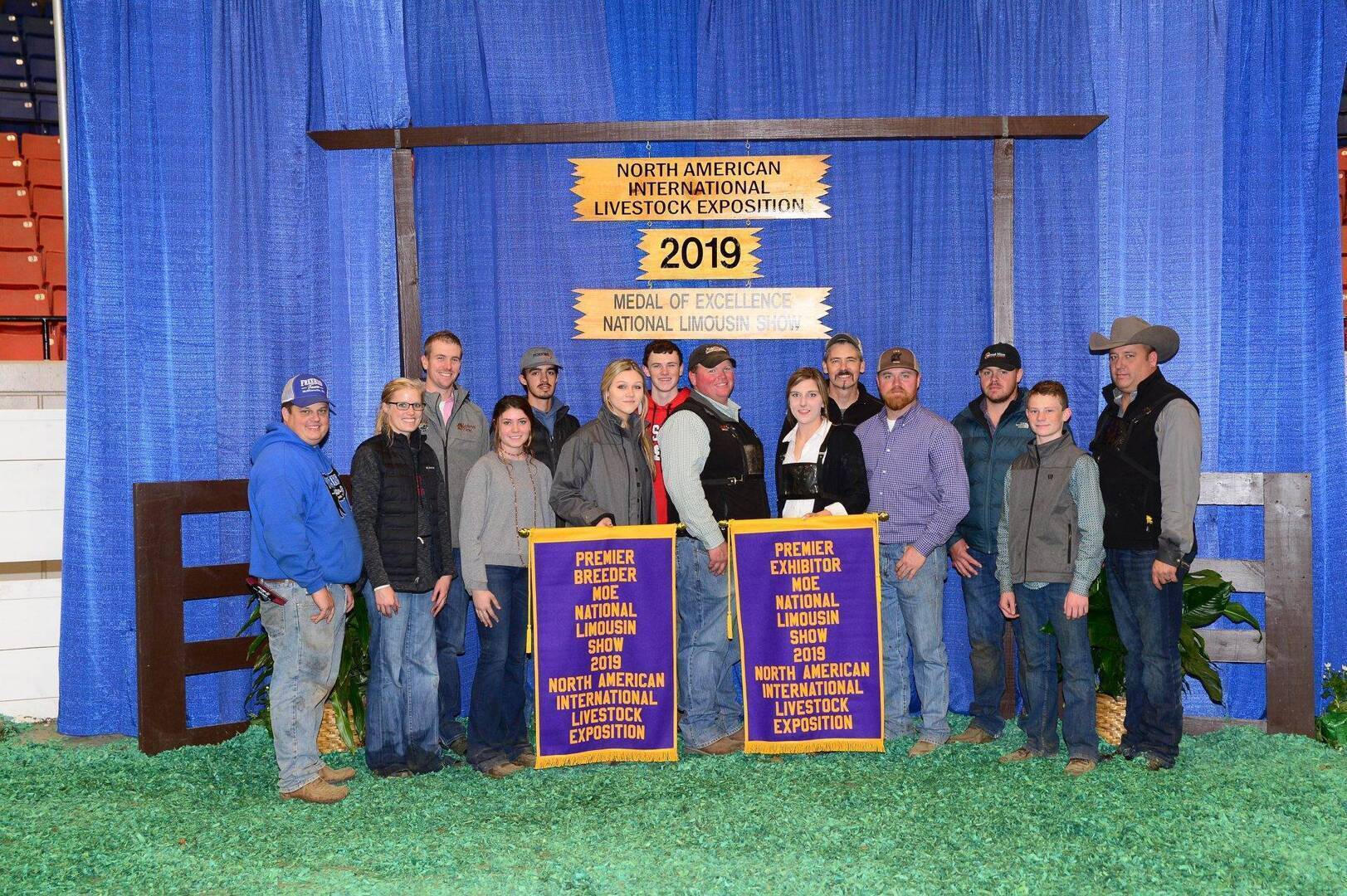 A group of people stands indoors on green shavings, posing with two large award banners that read “North American International Livestock Exposition 2019.” A wooden sign and blue curtain are in the background.