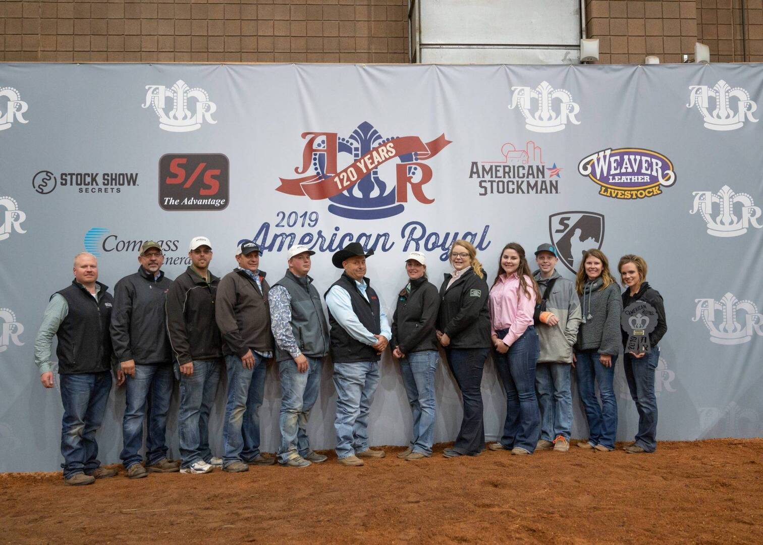 A group of people in jackets and jeans stand in a row on reddish dirt in front of a large “2019 American Royal” banner featuring sponsor logos and a “120 Years” ribbon.