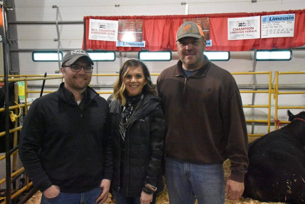 Three people stand smiling indoors next to cattle pens at a livestock event, with red banners for Limousin bulls and females hanging behind them. They are dressed in casual, farm-appropriate clothing.