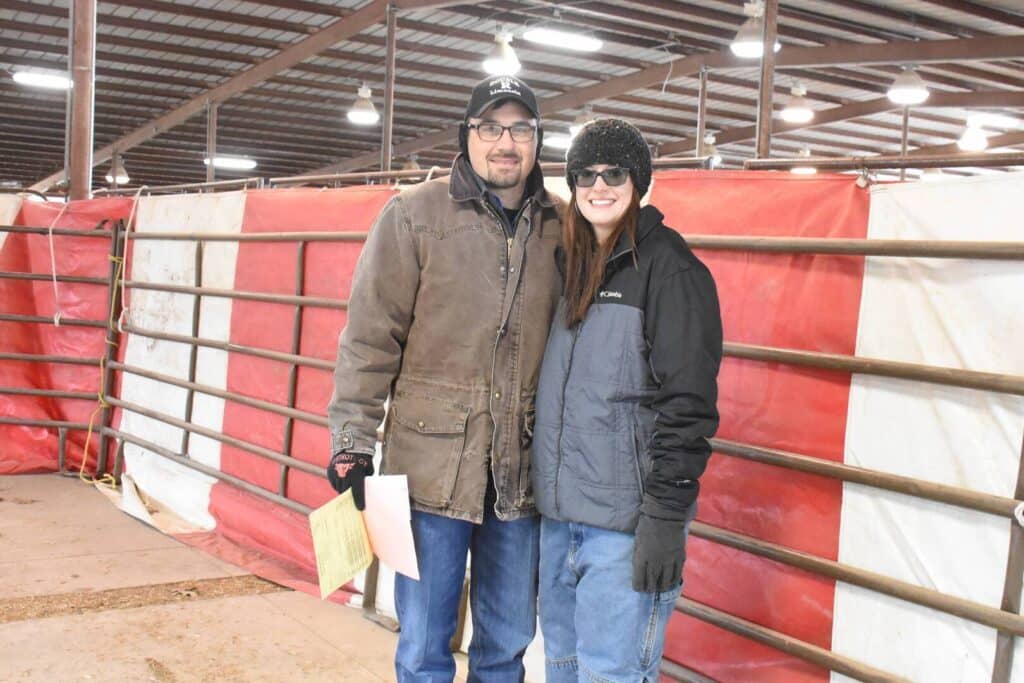 Two people wearing winter jackets, gloves, and hats stand smiling together inside a large, covered livestock arena with metal railings and a red and white barrier in the background.