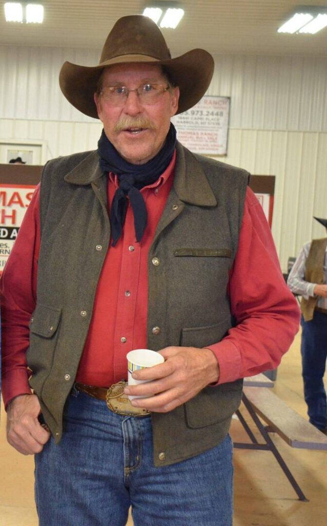 A man wearing a cowboy hat, glasses, red shirt, vest, and blue jeans holds a paper cup and smiles indoors, standing in front of a bench with signs and another person in the background.