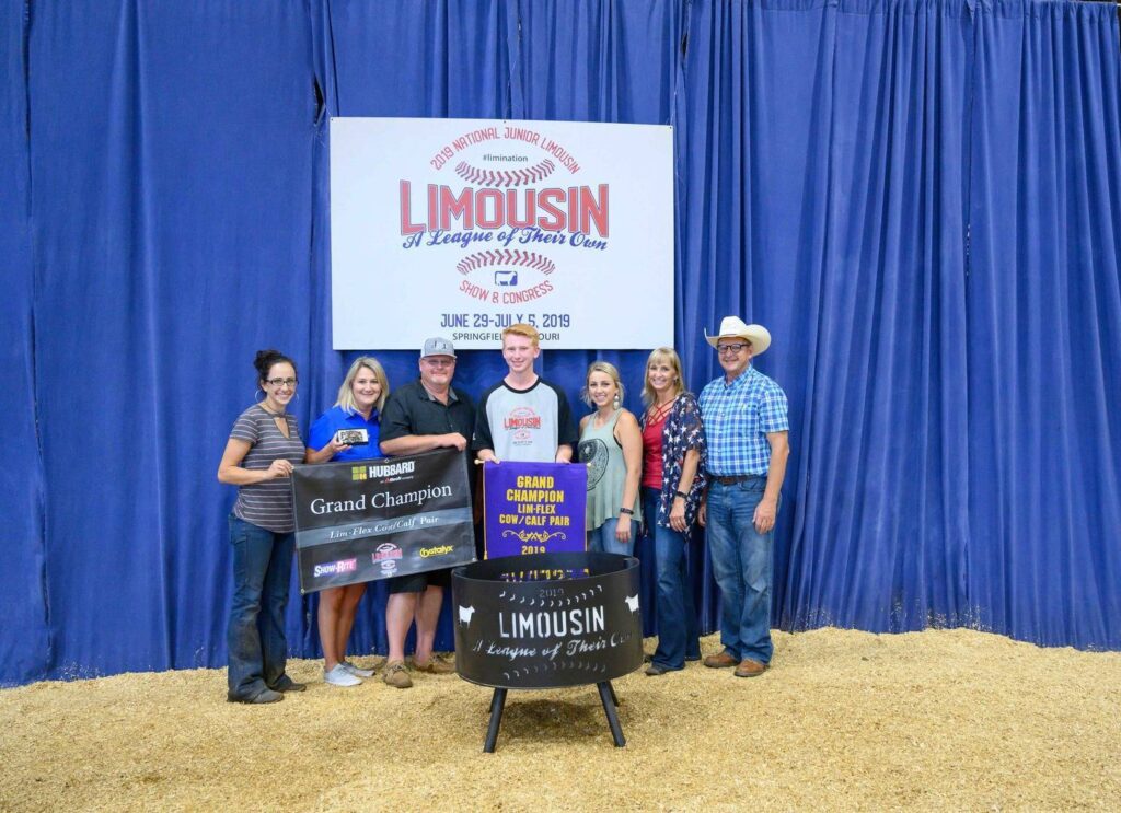 Seven people stand in front of blue curtains and a Limousin Grand Champion banner, holding awards and a sign, at a livestock event. A trophy and decorated table are in front of the group.