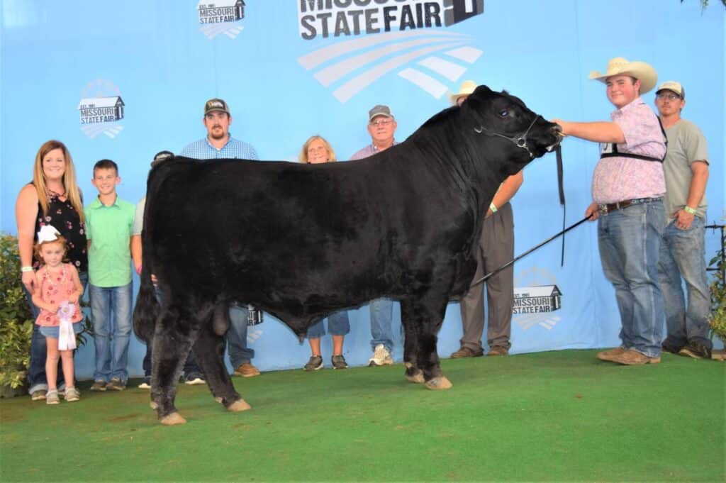 A group of people pose with a large black bull on green turf in front of a blue Missouri State Fair backdrop. Two men hold the bull, while others, including children, stand nearby and smile.