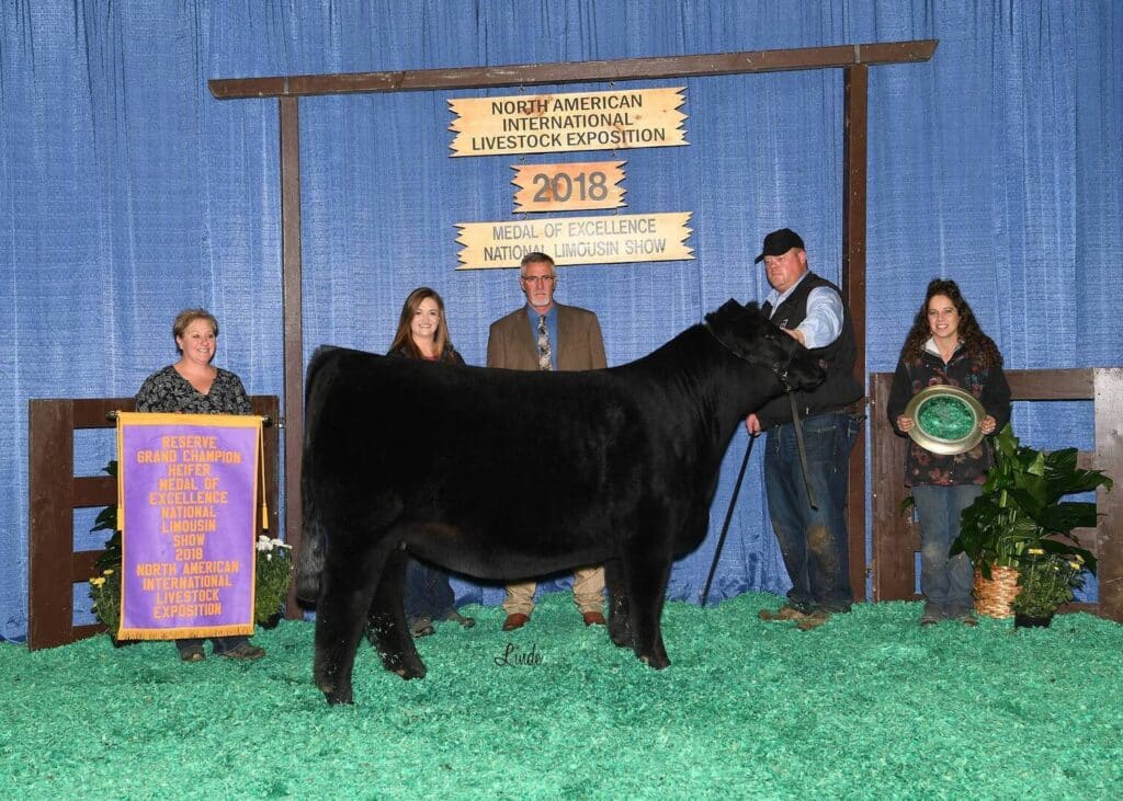 Four people pose with a black cow at the 2018 North American International Livestock Exposition. One person holds a purple award banner, another holds a green plate, and blue curtains are in the background.