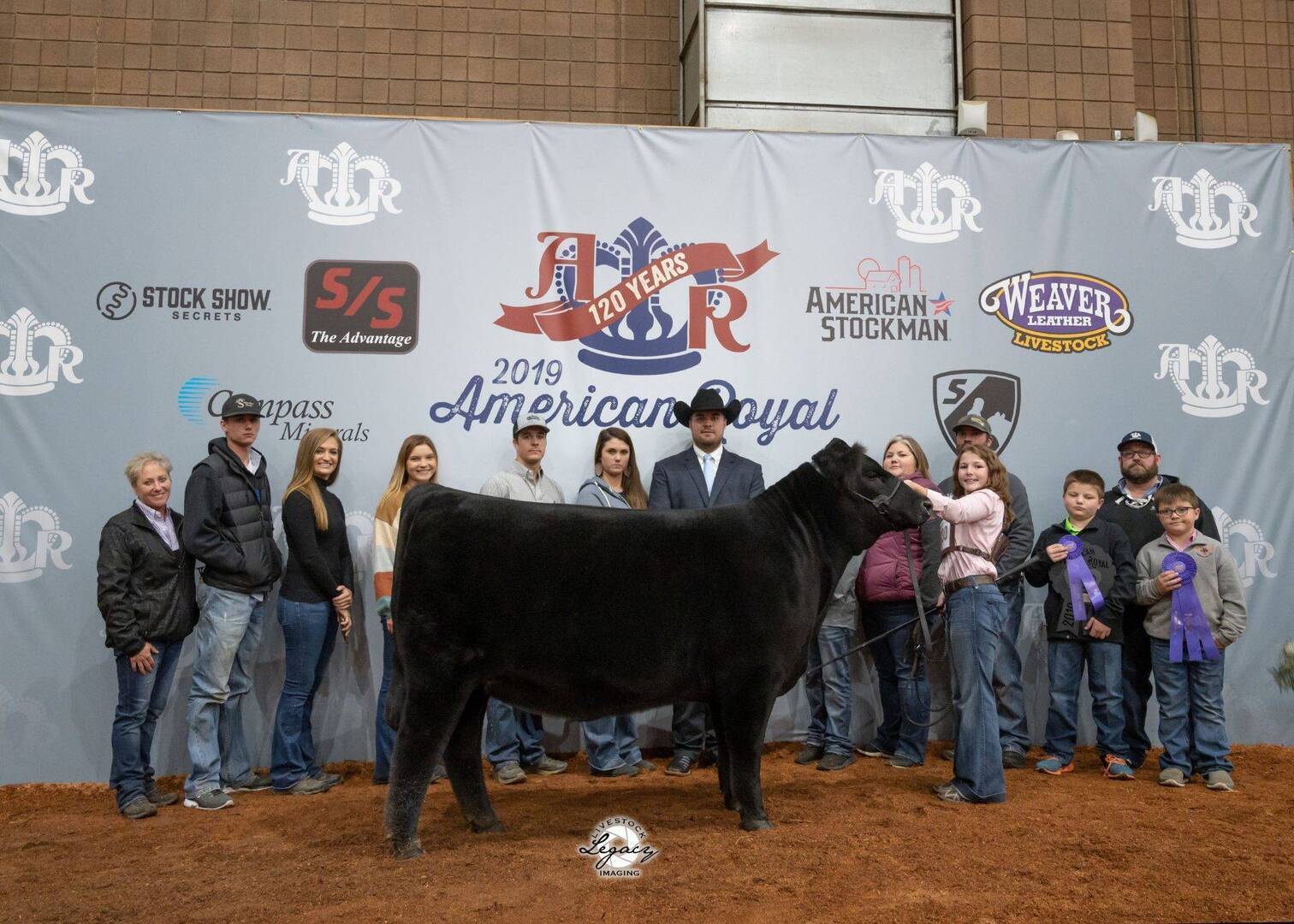 A group of people stand behind a black cow and its handler at the 2019 American Royal livestock show, posing for a photo in front of a banner with sponsor logos and event branding. Some hold ribbons and awards.