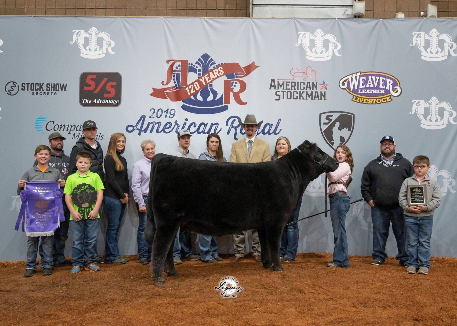 A group of people, including children, stand behind a black cow in a show ring. They pose with banners, ribbons, and a plaque in front of a backdrop reading 2019 American Royal with various sponsor logos.