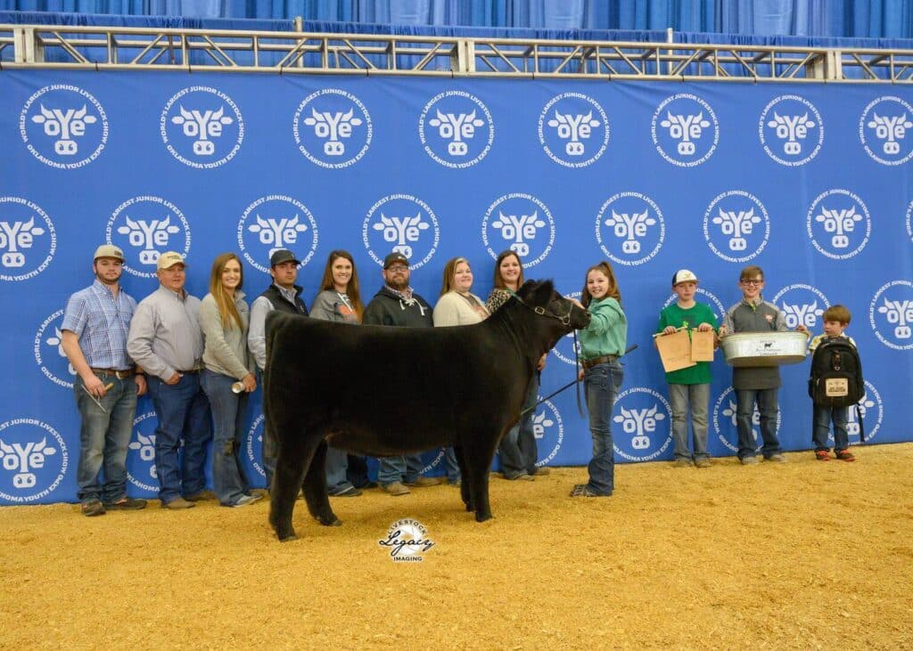 A group of people pose indoors with a black cow in front of a blue backdrop featuring OYE logos. Several kids hold awards, and everyone stands on a straw-covered floor.