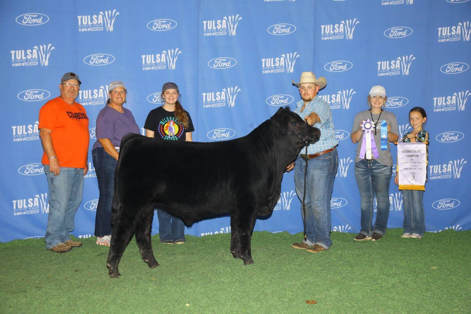 A group of people stand with a black cow in front of a blue Tulsa State Fair backdrop; two hold a ribbon and banner, suggesting the cow has won a prize.