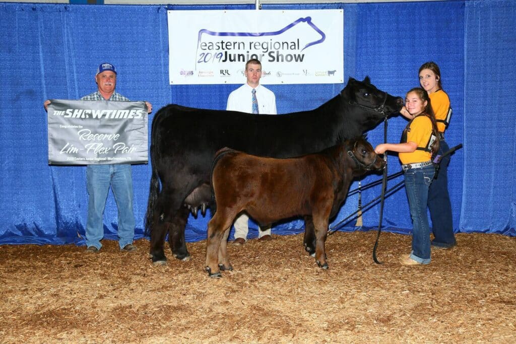 Four people stand with a black cow and a calf at the Eastern Regional Junior Show. One person holds a banner reading “Reserve Lim Flex Pair.” Blue curtains and a show sign are in the background.
