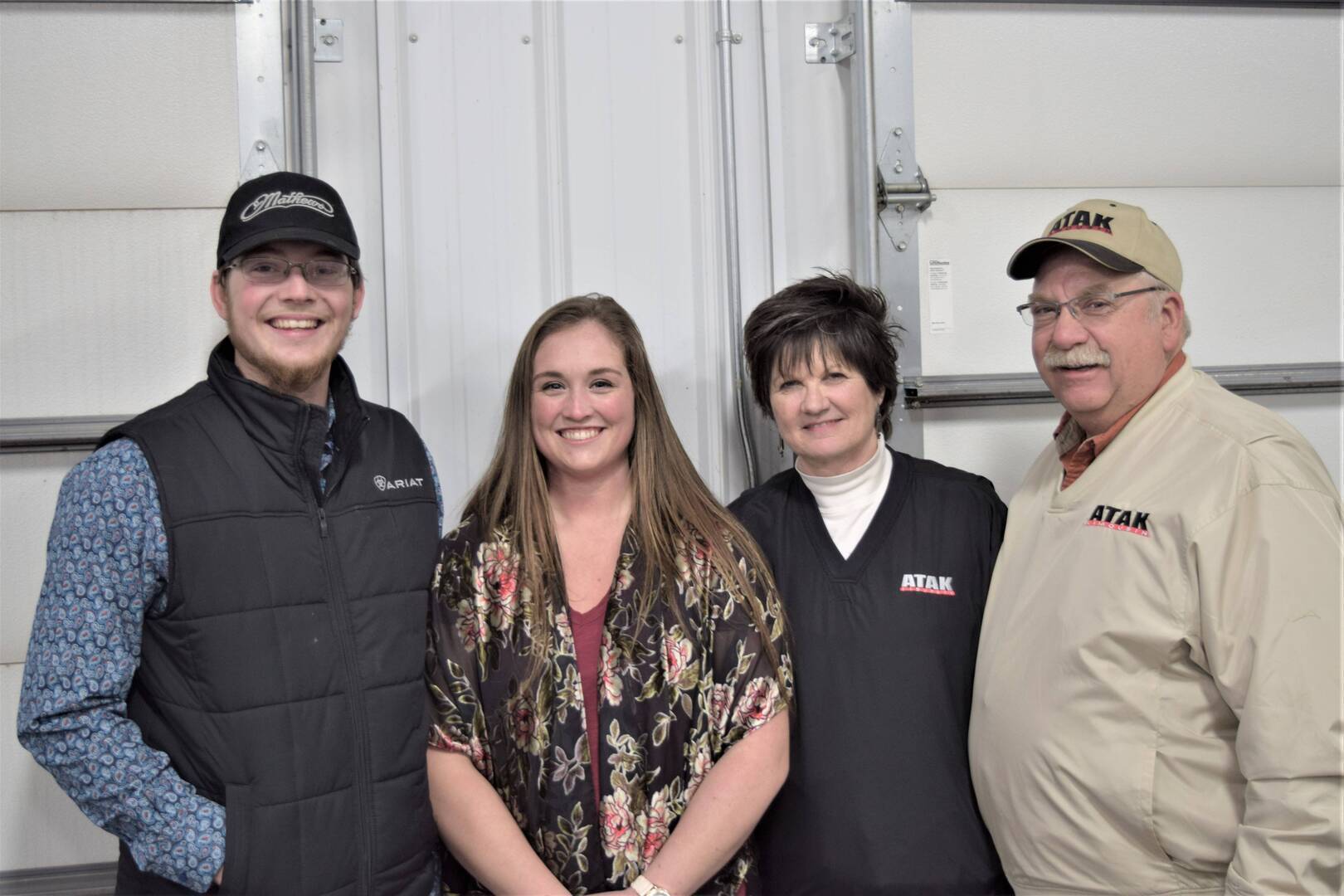 Four people stand side by side indoors, smiling at the camera. Two men wear hats and jackets with ATAK logos, while two women stand between them, dressed in casual clothing. They are in front of a light-colored wall.