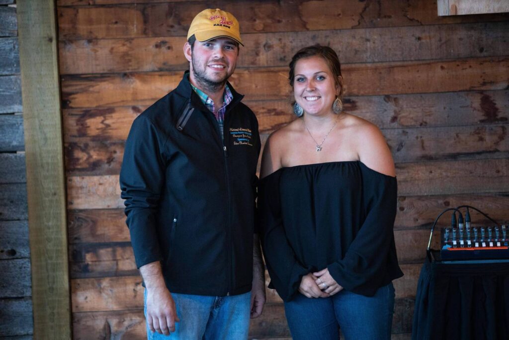 A man in a yellow cap and black jacket stands next to a woman in an off-the-shoulder black top and earrings. They are smiling and posing in front of a rustic wooden wall.