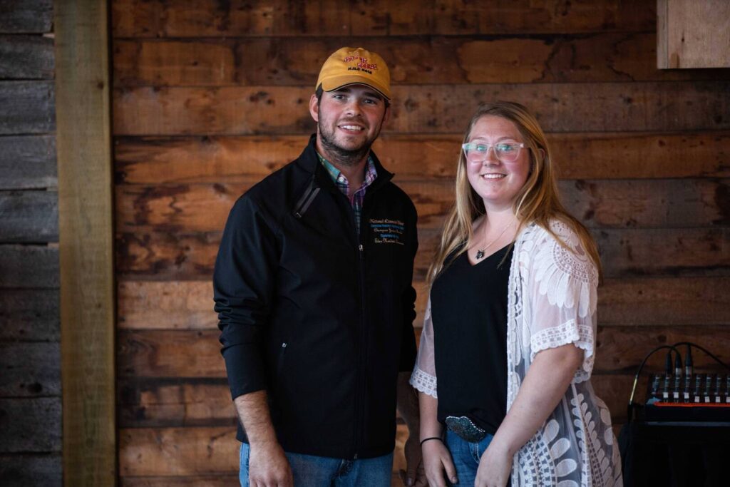 A man in a yellow hat and black jacket stands next to a woman in glasses and a white lace cardigan, both smiling, in front of a rustic wooden wall.