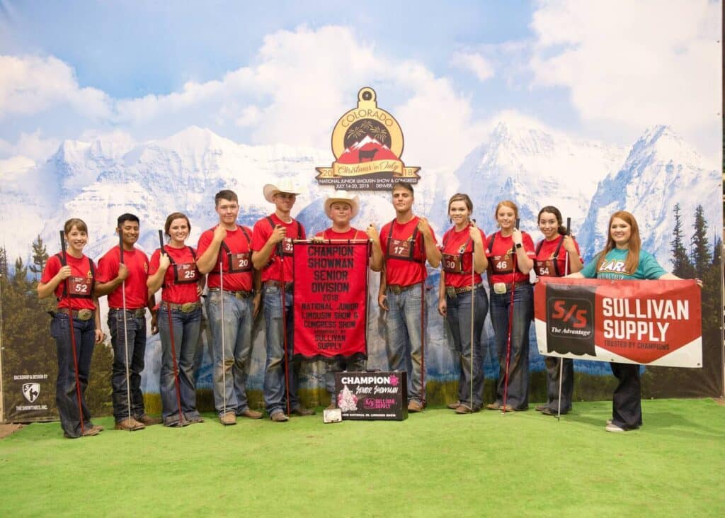 A group of ten young people in red shirts and jeans stand indoors in front of a mountain backdrop, holding awards and banners, including one reading “Champion Showman Senior Division.” Two adults stand on either side.