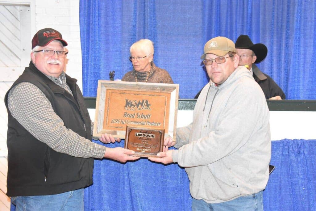 Two men in casual attire smile and hold a framed award that reads “ISWA Brad Schutt 2020 Limousin Producer,” while two people stand at a podium in the background with a blue curtain behind them.